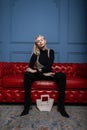 Stylish young woman in fashionable black clothes with a white pearl bag in a vintage pearl top sits on a leather red sofa on blue Royalty Free Stock Photo