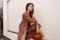 Stylish young woman in fashionable coat in fashion beige pants with brown leather handbag in shoe straightens long hair near Royalty Free Stock Photo