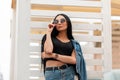 Stylish young woman model with black long hair in fashionable jeans clothes straightens trendy sunglasses. Attractive girl hipster Royalty Free Stock Photo