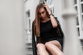 Stylish young woman model in an elegant short black dress in a fashionable black leather jacket sits on a vintage staircase near a Royalty Free Stock Photo