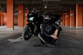 Stylish young woman motorcycle rider with beautiful eyes in black protective gear and full-face helmet near her bike on Royalty Free Stock Photo