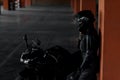 Stylish young woman motorcycle rider in black protective gear and full-face helmet near her bike on underground parking. Royalty Free Stock Photo