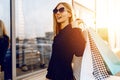 Stylish young woman in sunglasses and dress, with shopping bags, in the city against the background of a shopping center Royalty Free Stock Photo