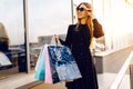 Stylish young woman in sunglasses and dress, with shopping bags, in the city against the background of a shopping center Royalty Free Stock Photo