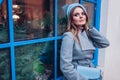 Stylish young woman wearing coat blue beret holding small purse sitting by cafe window outdoors. Autumn fashion Royalty Free Stock Photo