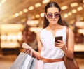 Stylish young woman wearing sunglasses using a cell phone while carrying bags during a shopping spree. Young brunette Royalty Free Stock Photo