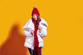 Stylish young woman in a white down coat and knite red hat on yellow background