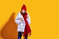 Stylish young woman in a white down coat and knite red hat on yellow background