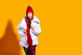 Stylish young woman in a white down coat and knite red hat on yellow background