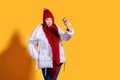 Stylish young woman in a white down coat and knite red hat on yellow background