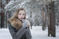 A stylishly dressed, beautiful girl in a gray coat with a fur collar stands alone in a winter forest, warming her hands in fur mit