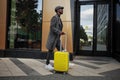 Stylishly dressed male model walks down city street with yellow suitcase in hand. Black man tourist outdoors near hotel Royalty Free Stock Photo