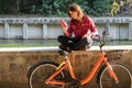 Subject ecological bicycle transport. Young Caucasian woman in shirt student sits resting in a park near the lake for rent orange Royalty Free Stock Photo
