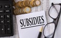 SUBSIDIES - word on a white piece of paper on the background of a calculator, pennies and glasses