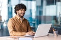 Succesful arab man with beard sitting on chair by desk with laptop and tablet next to glass walls. Portrait of young Royalty Free Stock Photo