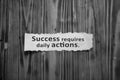 Success requires daily actions. Inspirational words on white torn paper note on wooden wall background in black and white. Royalty Free Stock Photo