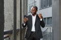 Successful african american boss outside office building using phone, businessman in suit celebrating victory Royalty Free Stock Photo