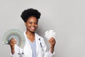 Successful african american woman doctor gp smiles and holds fan of money and packs of pills against grey background