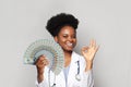 Successful african american woman doctor smiles and shows OK and holds fan of moneyagainst white background