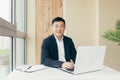 Successful Asian businessman, in a black business suit, works on a laptop, relaxes in a stylish office Royalty Free Stock Photo