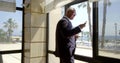 Successful businessman in black suit is drinking coffee near big window with sea view, using smartphone