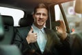 Successful businessman making online payment using bank card while riding on back seat of car showing like sign, looks satisfied, Royalty Free Stock Photo