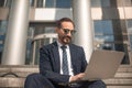 Successful businessman works on laptop sitting on steps of business center. Smiling man wearing business suit and Royalty Free Stock Photo