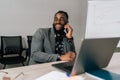 Successful cheerful African businessman in suit talking on smartphone sitting at office desk with laptop. Happy Royalty Free Stock Photo