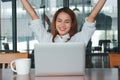 Successful cheerful young Asian businesswoman with laptop raising hands in office. Thinking and thoughtful business concept. Royalty Free Stock Photo