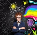 Successful clever child boy with ginger hair, glasses and crossed arms on lightbulb, science and arts scetch background. Royalty Free Stock Photo
