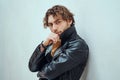 Daring and cool male model with curly hair posing in leather coat in a bright studio and leaning on the wall