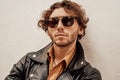 Portrait of a handsome guy with curly hair posing in the bright studio wearing sunglasses and leather coat Royalty Free Stock Photo