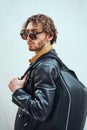Portrait of a handsome guy with curly hair posing in the bright studio wearing sunglasses, leather coat and a backpack Royalty Free Stock Photo