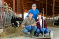 Successful elderly dairy farm owner with son and teen grandson standing in stall with cows
