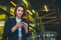 Successful female banker using smartphone outdoors while standing near his office background yellow neon lights, young woman