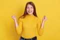 Successful female being happy, clenches fists, accomplishes goal, exclaims, stands amused over yellow background, looks at camera