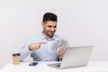 Successful happy elegant businessman sitting in office workplace pointing money dollars and smiling at laptop Royalty Free Stock Photo