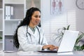 Successful hispanic female doctor working inside medical office at desk, woman typing on laptop, smiling with video call Royalty Free Stock Photo