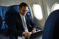 Successful millennial businessman in black suit using laptop while sitting in airplane cabin