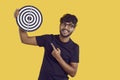 Successful positive indian male freelancer demonstrates darts board while on yellow background.