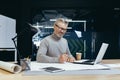 Successful senior architect at work in office, mature gray haired man drawing blueprints on layout and plan smiling and Royalty Free Stock Photo