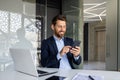 A successful and smiling young male businessman is sitting in the office at a desk in a suit and using a mobile phone Royalty Free Stock Photo