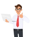 Successful young businessman holding laptop computer and making thumbs up gesture. Person showing good, agree or approval sign.