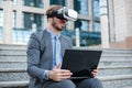 Successful young businessman using virtual reality simulator goggles and working on a laptop in front of an office building Royalty Free Stock Photo