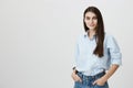 Successful young female blogger standing with hands in pocket and looking at camera with confident and pleasant look