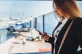 Successful young female freelancer standing near window at airport holding a phone texting