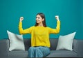 Successful young girl demonstrate strength and power raising arms showing biceps, proud of achievement, sitting on sofa