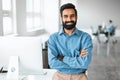 Successful young indian businessman in shirt standing with folded arms near desk, smiling at camera, office interior Royalty Free Stock Photo