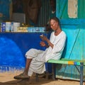 Sudanese man in traditional clothes sits on a bench in front of the shop for milk powder and listens to his mobile phone
