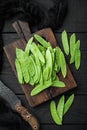 Sugar snap peas, raw ripe baby pods, on wooden cutting board, on black wooden background, top view flat lay